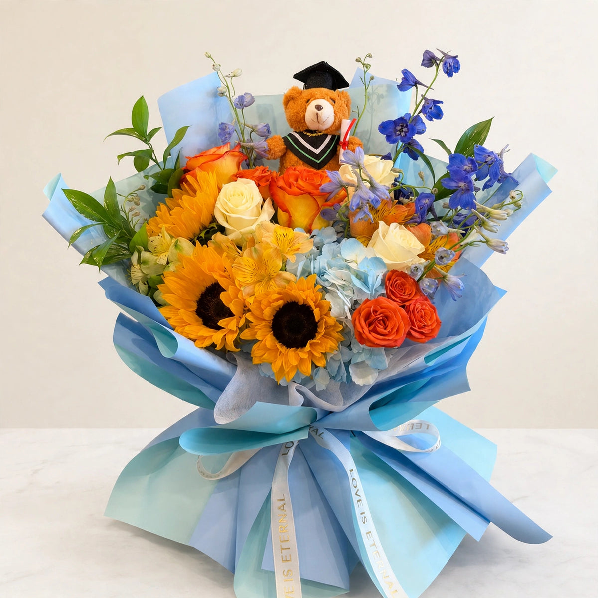 Sunshine Celebration Bouquet — sunflowers, orange and yellow roses and blue delphinium with a graduation teddy bear in a blue wrap, hand-arranged by LaLa Florist in Baldwin Park