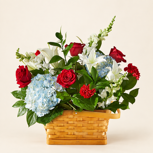 Patriotic sympathy basket with red roses, white lilies and blue hydrangeas arranged in a natural wicker basket, three-quarter view
