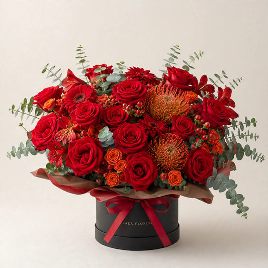 Crimson Prosperity Box — red roses, orange spray roses and pincushion protea in a black hat box by LaLa Florist in Baldwin Park