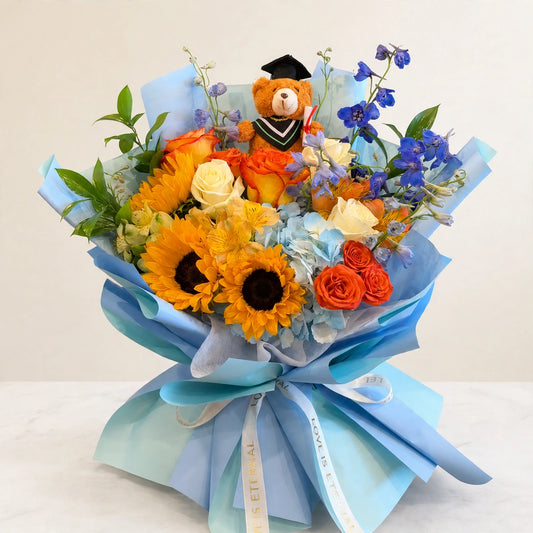 Sunshine Celebration Bouquet — sunflowers, orange and yellow roses and blue delphinium with a graduation teddy bear in a blue wrap, hand-arranged by LaLa Florist in Baldwin Park